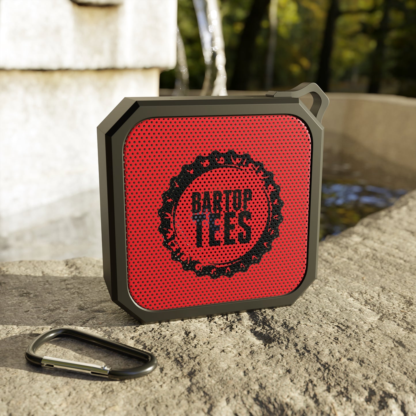 Compact red and black waterproof outdoor Bluetooth speaker with a carabiner clip resting on a stone ledge.