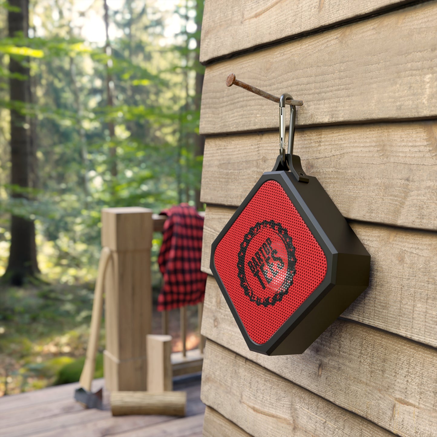Compact black and red outdoor Bluetooth speaker with a carabiner clip hanging on a rustic wooden wall.