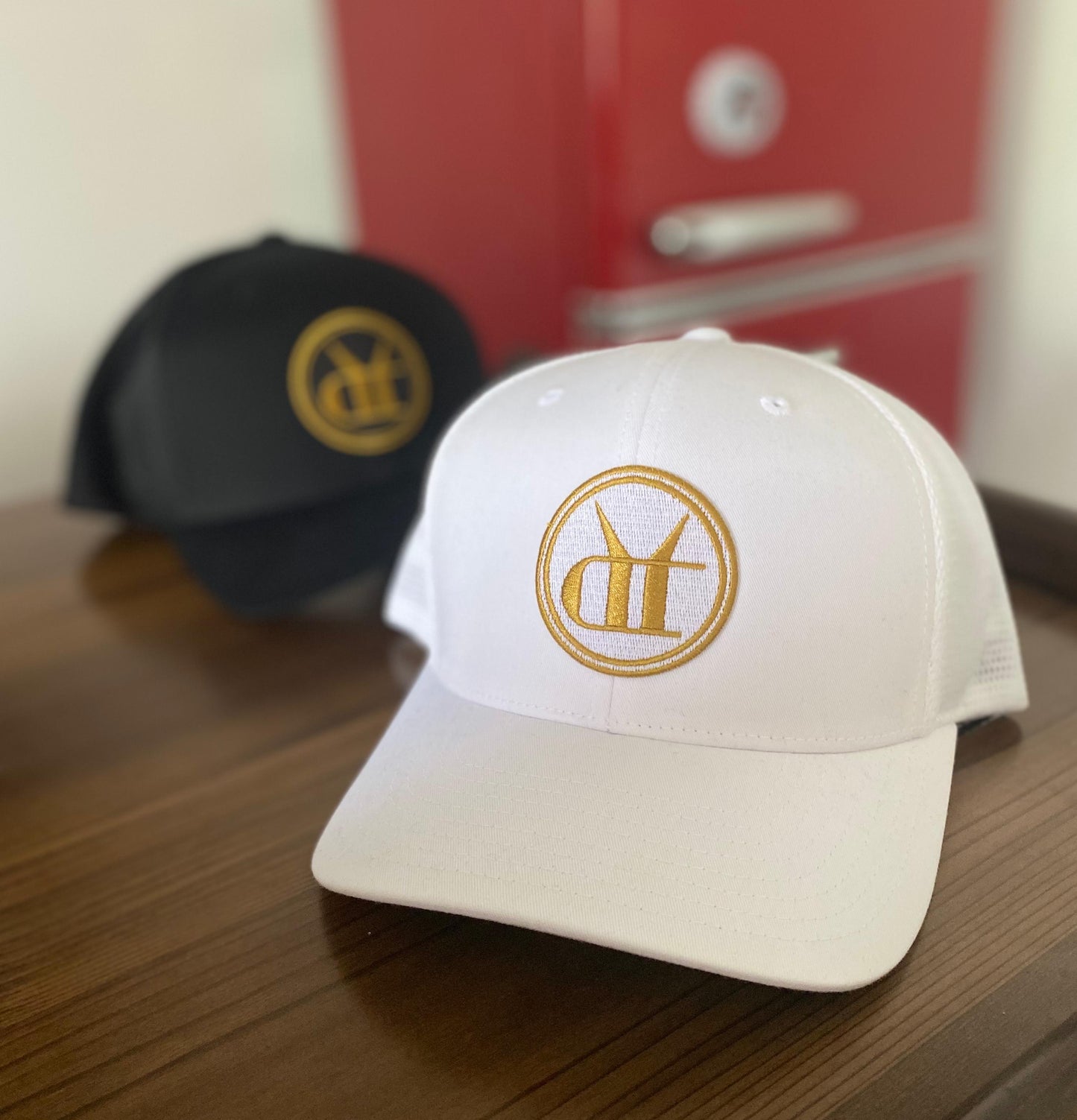 White and black snapback trucker hats featuring a circular gold embroidered Drink That logo on a wooden surface.