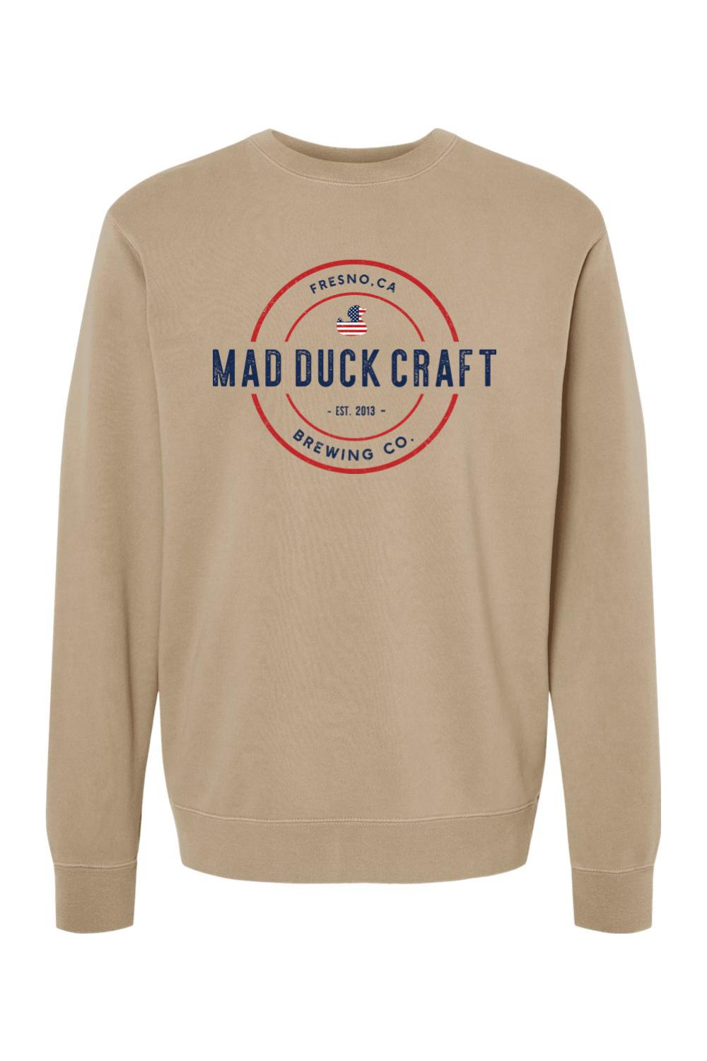 Tan pigment-dyed crewneck sweatshirt featuring a Mad Duck Craft Brewing logo with red, white, and blue American flag accents.
