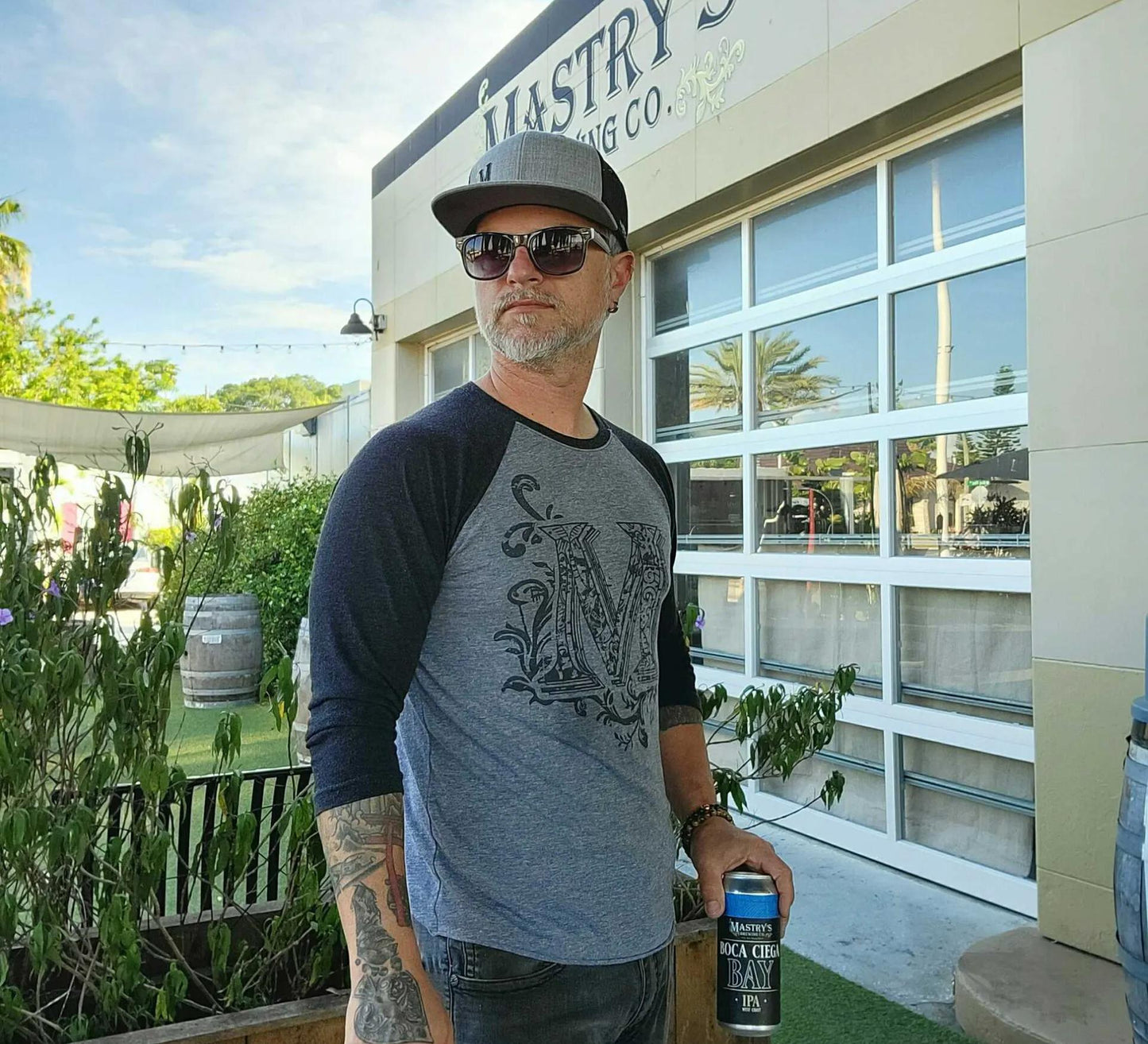 Man wearing a grey and navy blue raglan baseball shirt with a graphic M logo, standing outside Mastry’s Brewing Co.