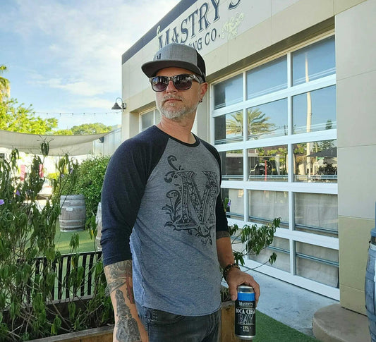 Man wearing a grey and navy blue raglan baseball shirt with a graphic M logo, standing outside Mastry’s Brewing Co.