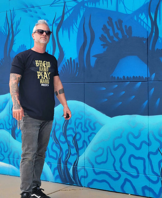 Man wearing a black Mastry’s Brewing Co. Brew Hard Play Hard graphic triblend t-shirt against a blue mural.
