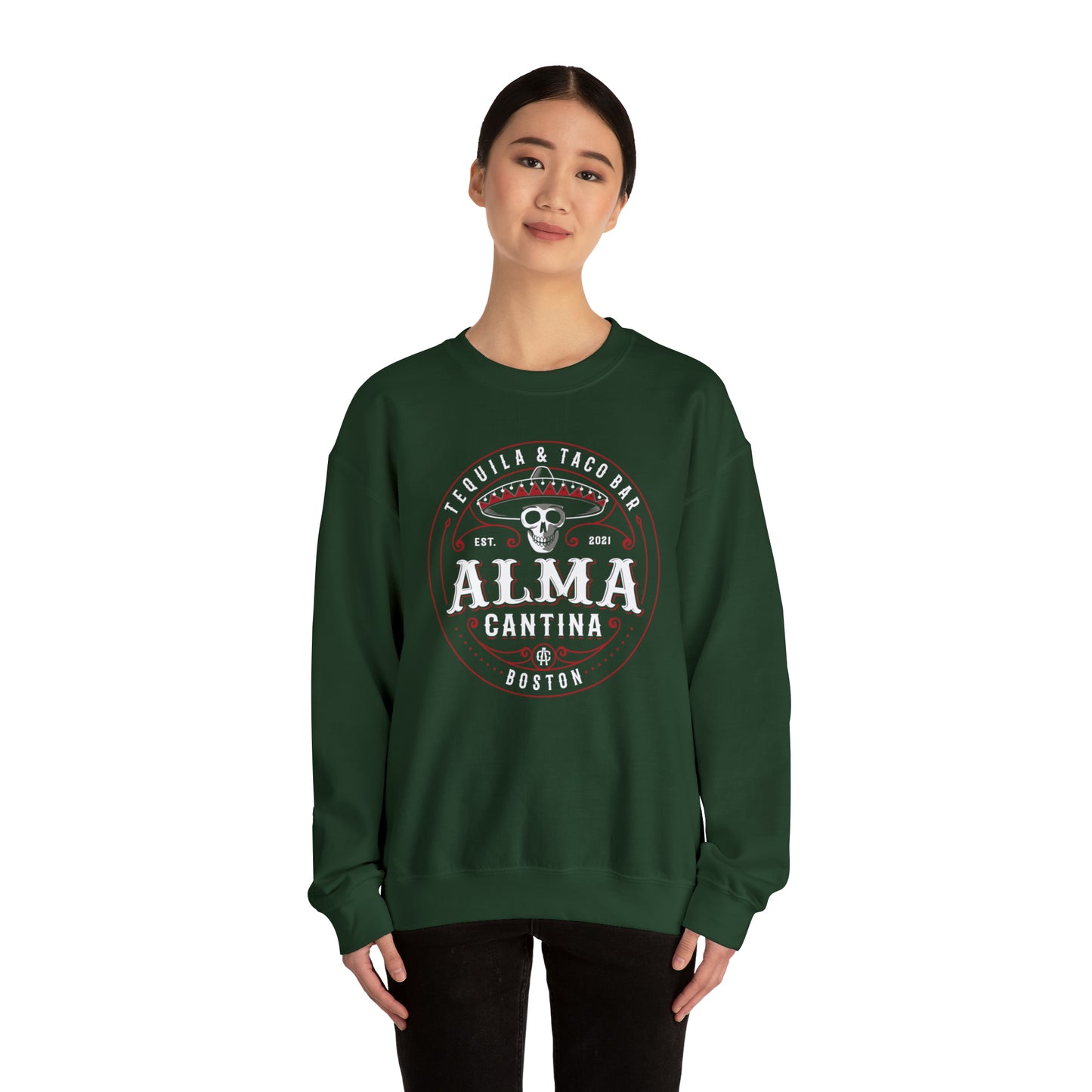 Woman wearing a dark green heavy blend crewneck sweatshirt featuring a white and red Alma Cantina skull logo.