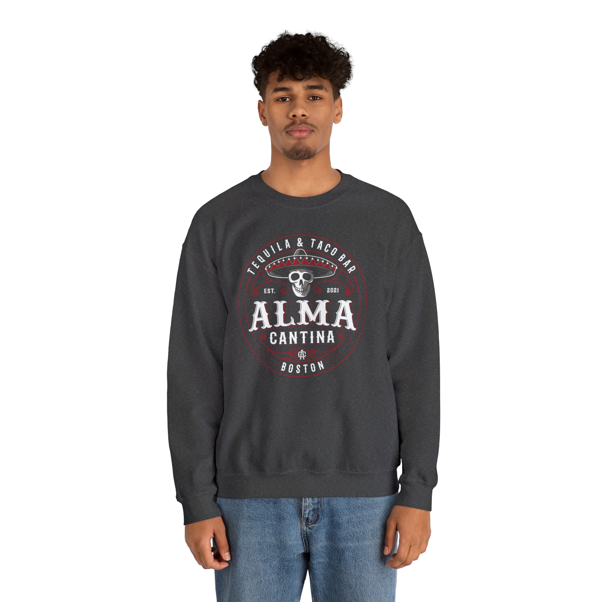 Man wearing a dark gray heavy blend crewneck sweatshirt with a circular Alma Cantina skull and sombrero graphic.