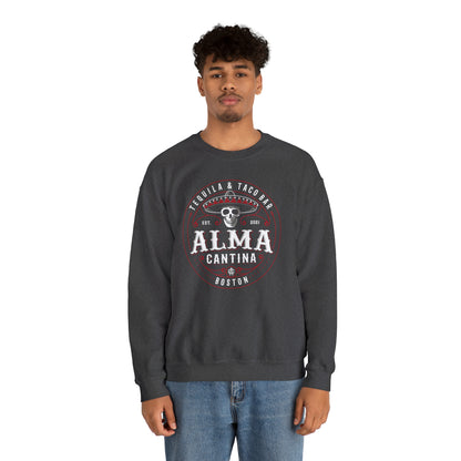Man wearing a dark gray heavy blend crewneck sweatshirt with a circular Alma Cantina skull and sombrero graphic.