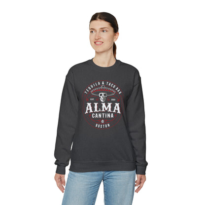 Woman wearing a dark gray heavy blend crewneck sweatshirt with a white and red Alma Cantina skull logo graphic.