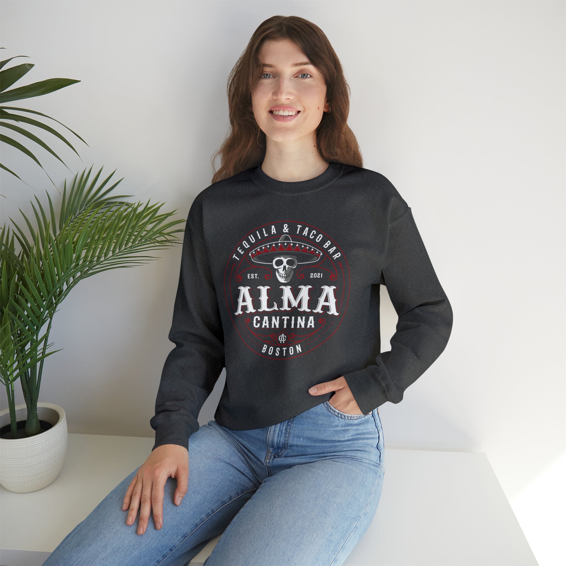 Woman wearing a charcoal grey heavy blend crewneck sweatshirt with a skull and Alma Cantina logo graphic.