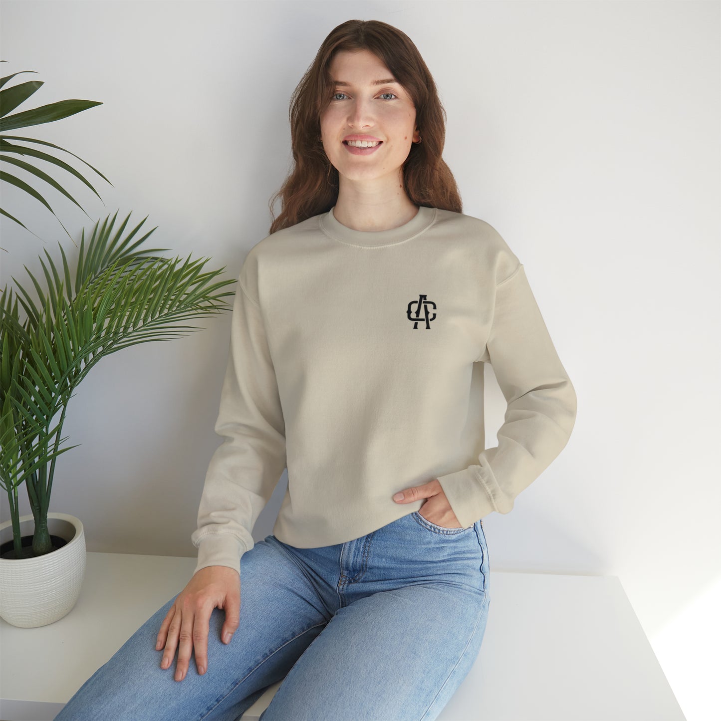 Woman wearing a sand-colored heavy blend crewneck sweatshirt with a black logo, paired with blue jeans.