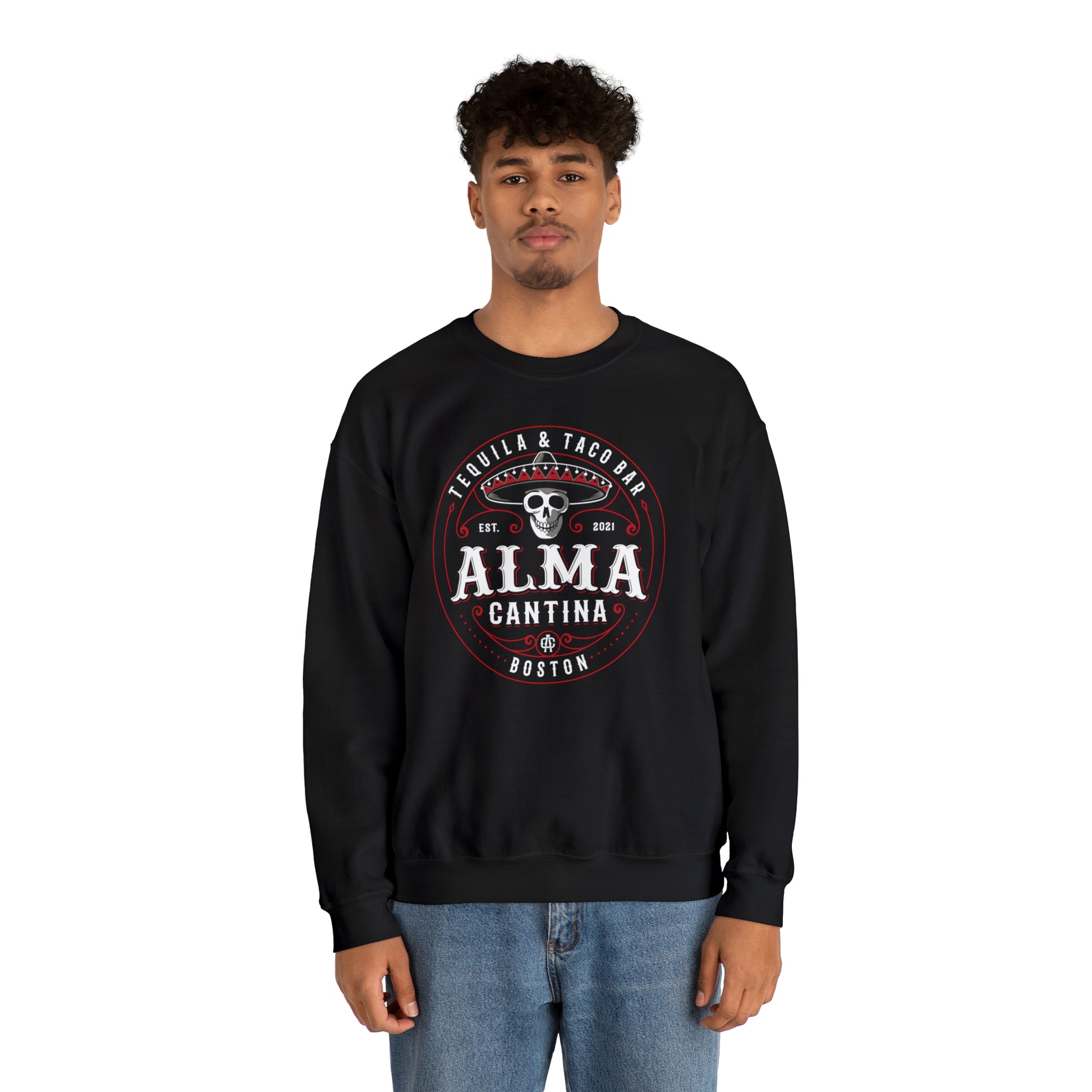 Man wearing a black heavy blend crewneck sweatshirt with a circular Alma Cantina skull and sombrero graphic.