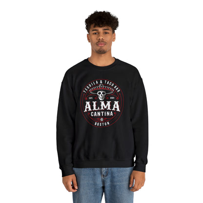 Man wearing a black heavy blend crewneck sweatshirt with a circular Alma Cantina skull and sombrero graphic.