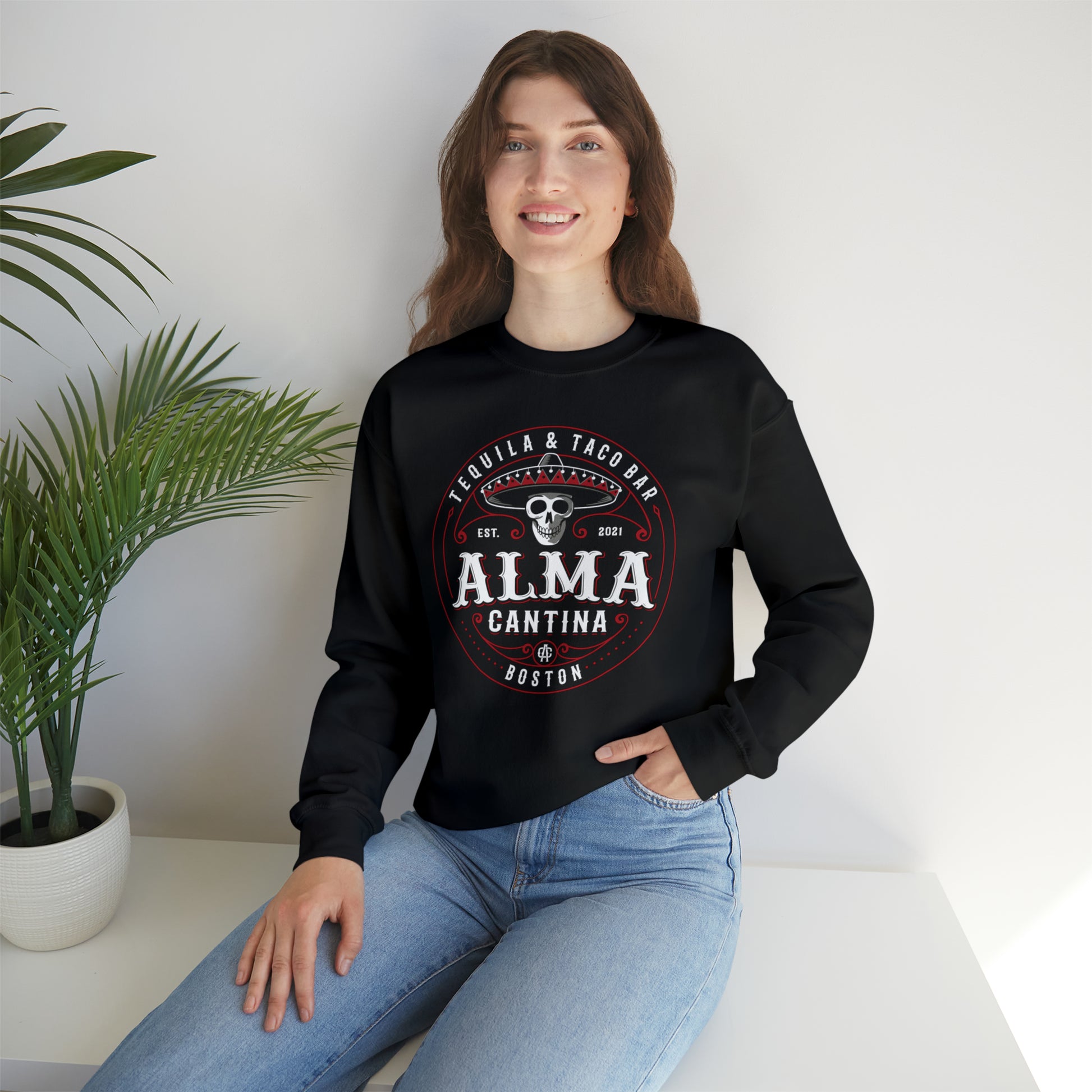 Woman wearing a black crewneck sweatshirt with a white and red Alma Cantina skull graphic, paired with blue jeans.