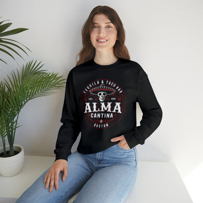 Woman wearing a black crewneck sweatshirt with a white and red Alma Cantina skull graphic, paired with blue jeans.