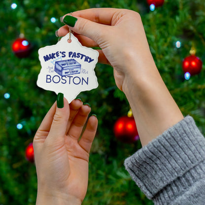 Glossy white ceramic ornament featuring a blue Mike's Pastry Boston design, held by hands against a Christmas tree.