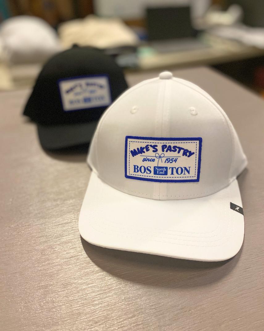 White and black structured snapback hats featuring an embroidered royal blue Mike's Pastry patch on the front.