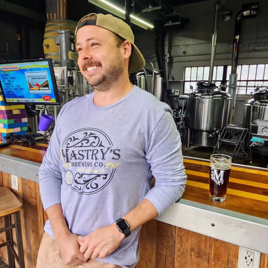 Man wearing a grey Mastry’s Brewing Co. long sleeve shirt with a circular logo in a brewery setting.