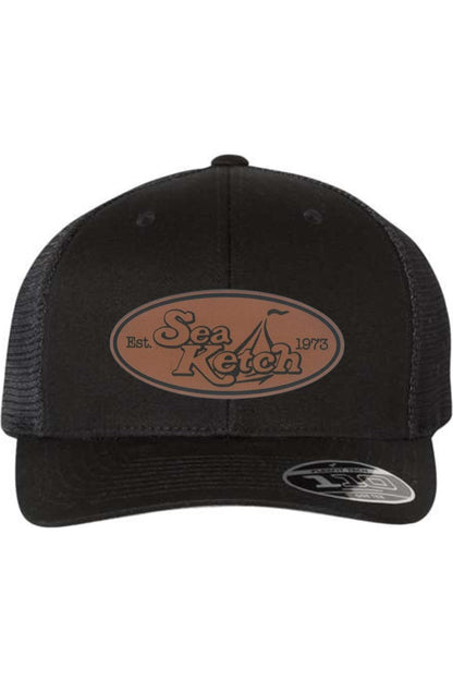 Black Flexfit 110 trucker cap with a brown oval leather patch featuring the Sea Ketch logo and mesh back.