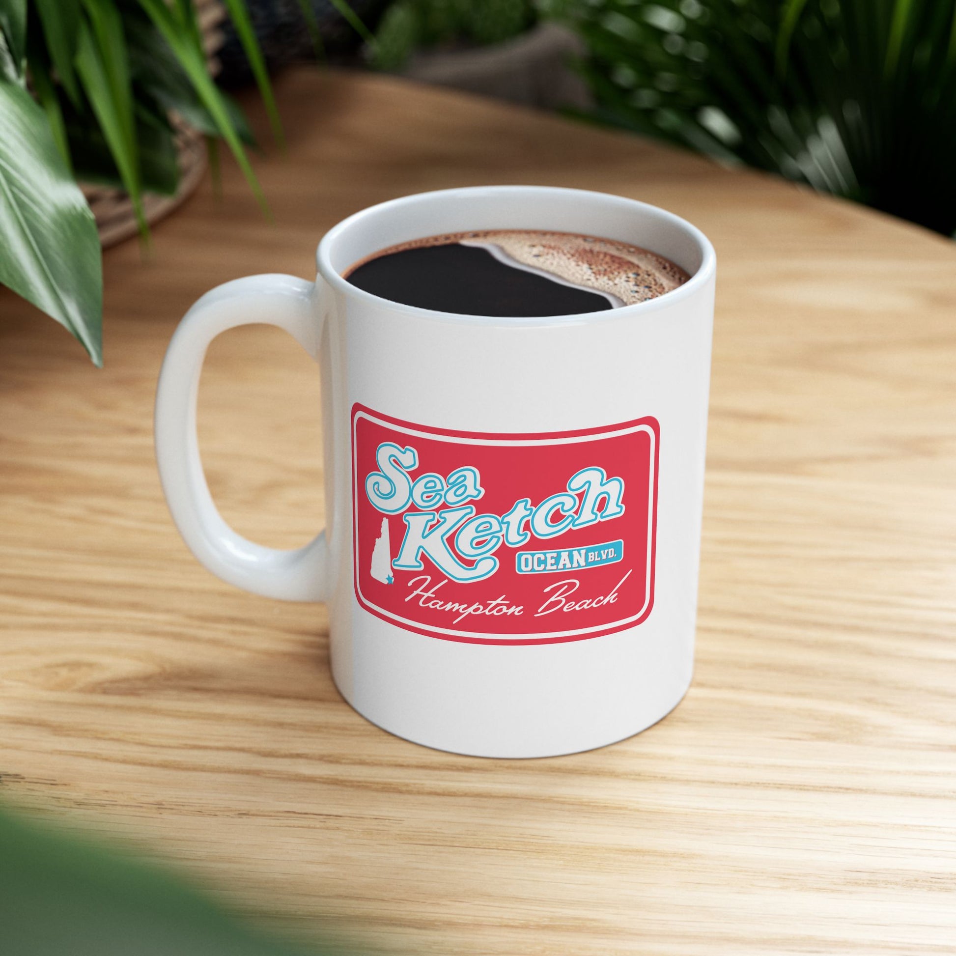 White ceramic mug featuring a red Sea Ketch Hampton Beach logo with a New Hampshire map on a wooden table.