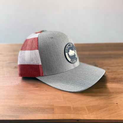 Heather gray trucker hat with American flag mesh back and White Bull Tavern PVC patch on a wooden surface.